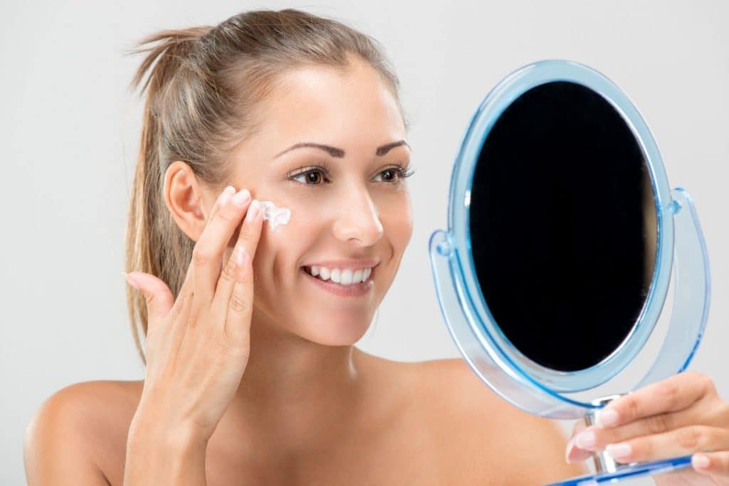 a smiling lady holding a mirror while putting a first aid beauty vegan cream