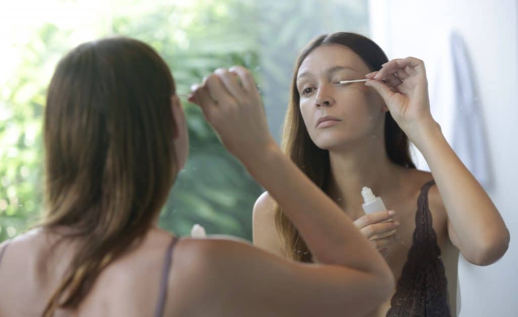 Young caucasian woman applying serum for eyebrows and eyelashes growth in front of the mirror. Night time beauty routine.
